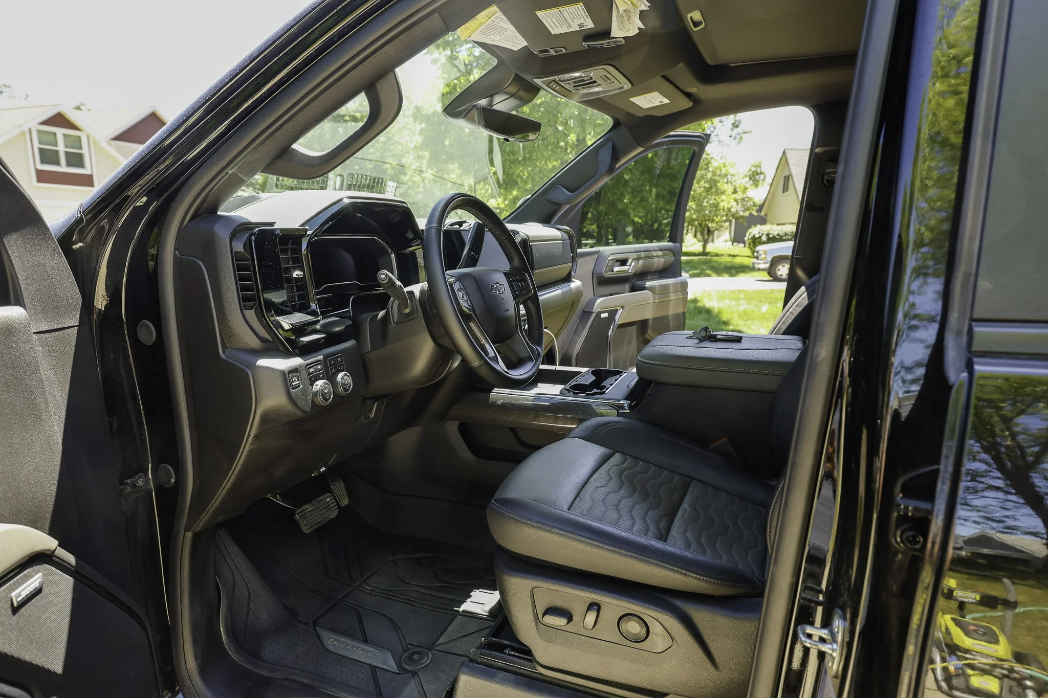 The interior of a Chevrolet pickup truck, showing the driver's seat, steering wheel, dashboard, and part of the center console, with a view of a residential neighborhood through the open door.