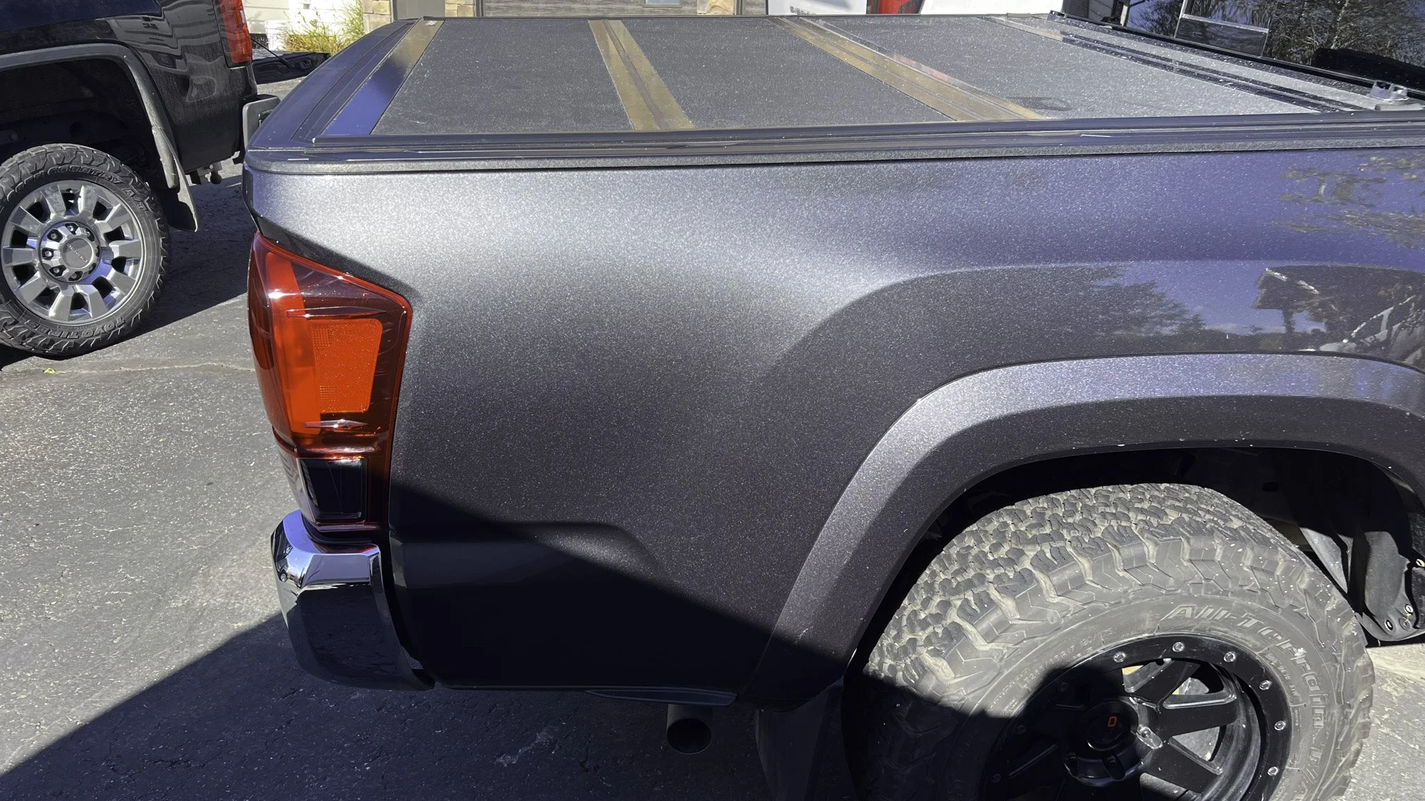 Close-up of the rear side of a dark gray pickup truck, showing the tail light, wheel with tire, and rear bumper, parked on an asphalt surface.