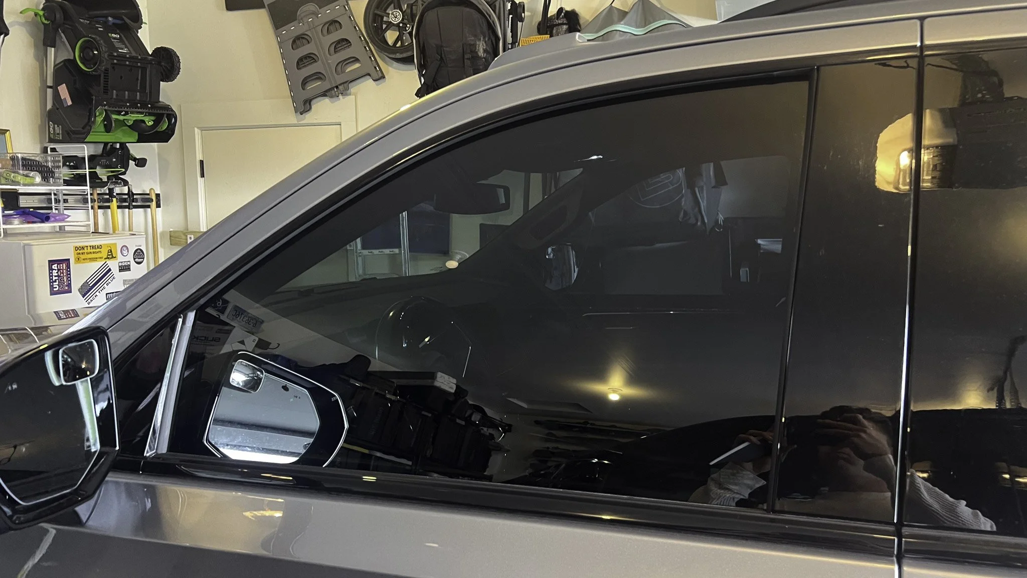 Close-up of a black car's side window reflecting the person taking the photo and the garage interior.