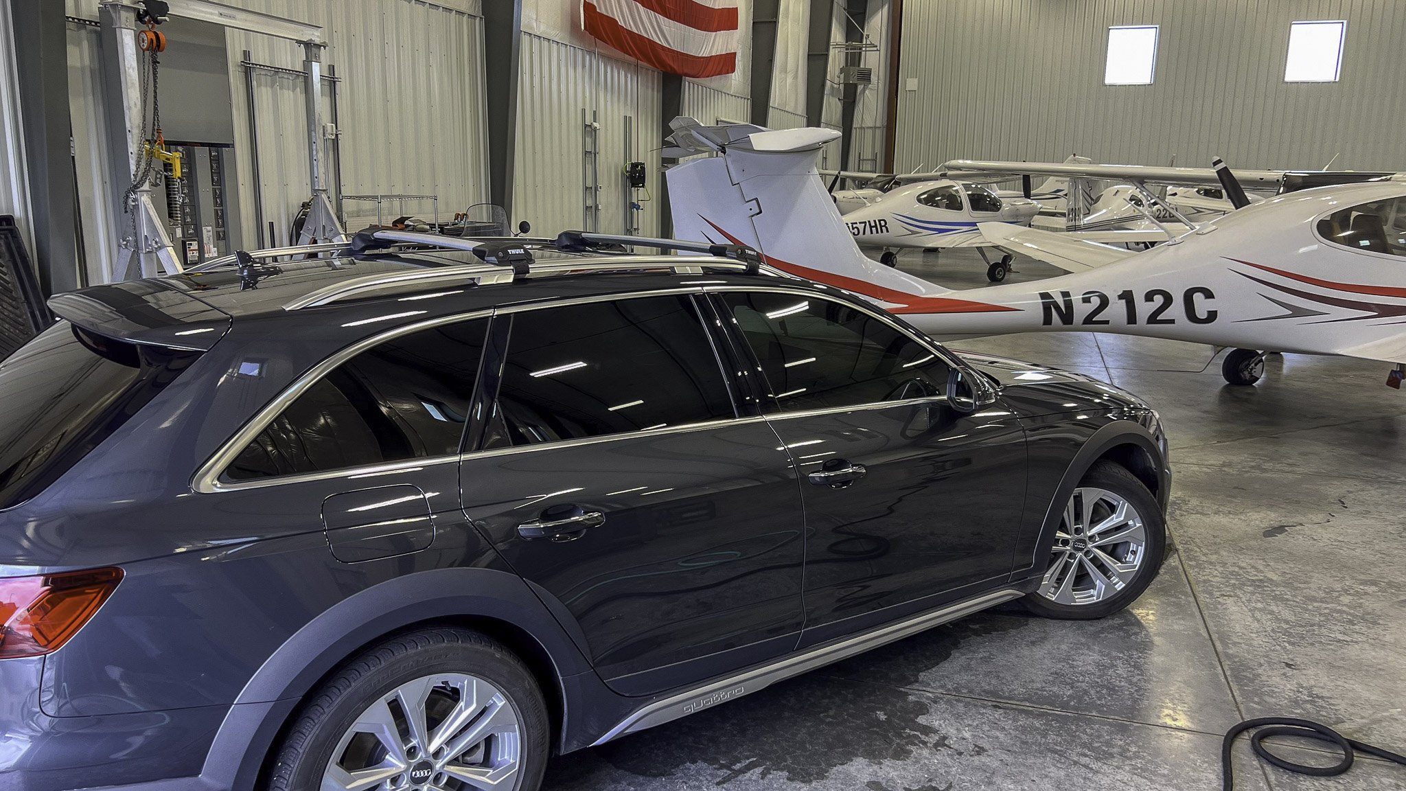Inside a hangar with a black Audi station wagon in the foreground and multiple small aircraft, including a white aircraft with red and gray stripes and the registration N212C, in the background.