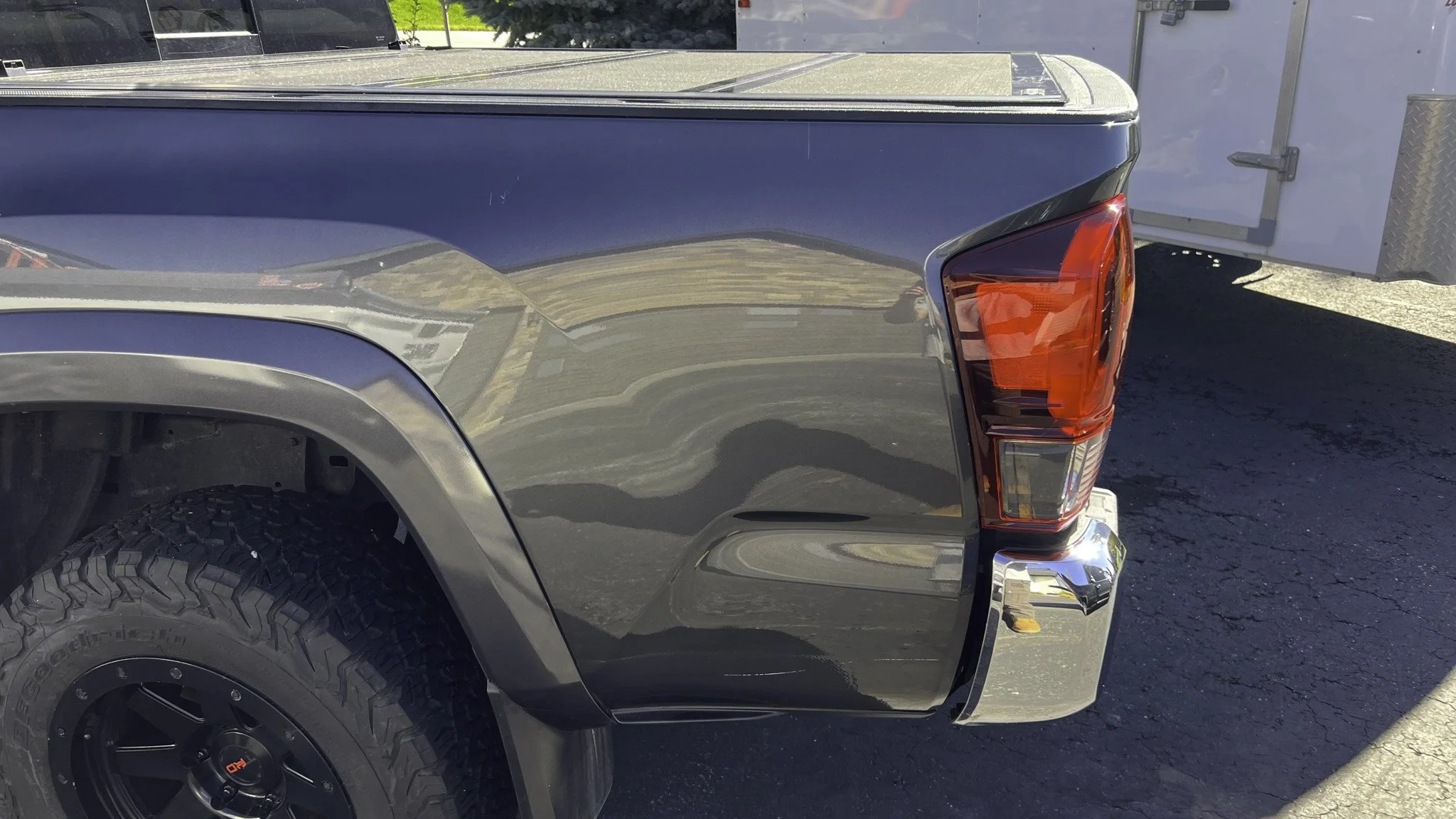 Rear right side of a black pickup truck with a large tire, red and clear taillight, chrome bumper, and a shiny exterior that reflects the surroundings.