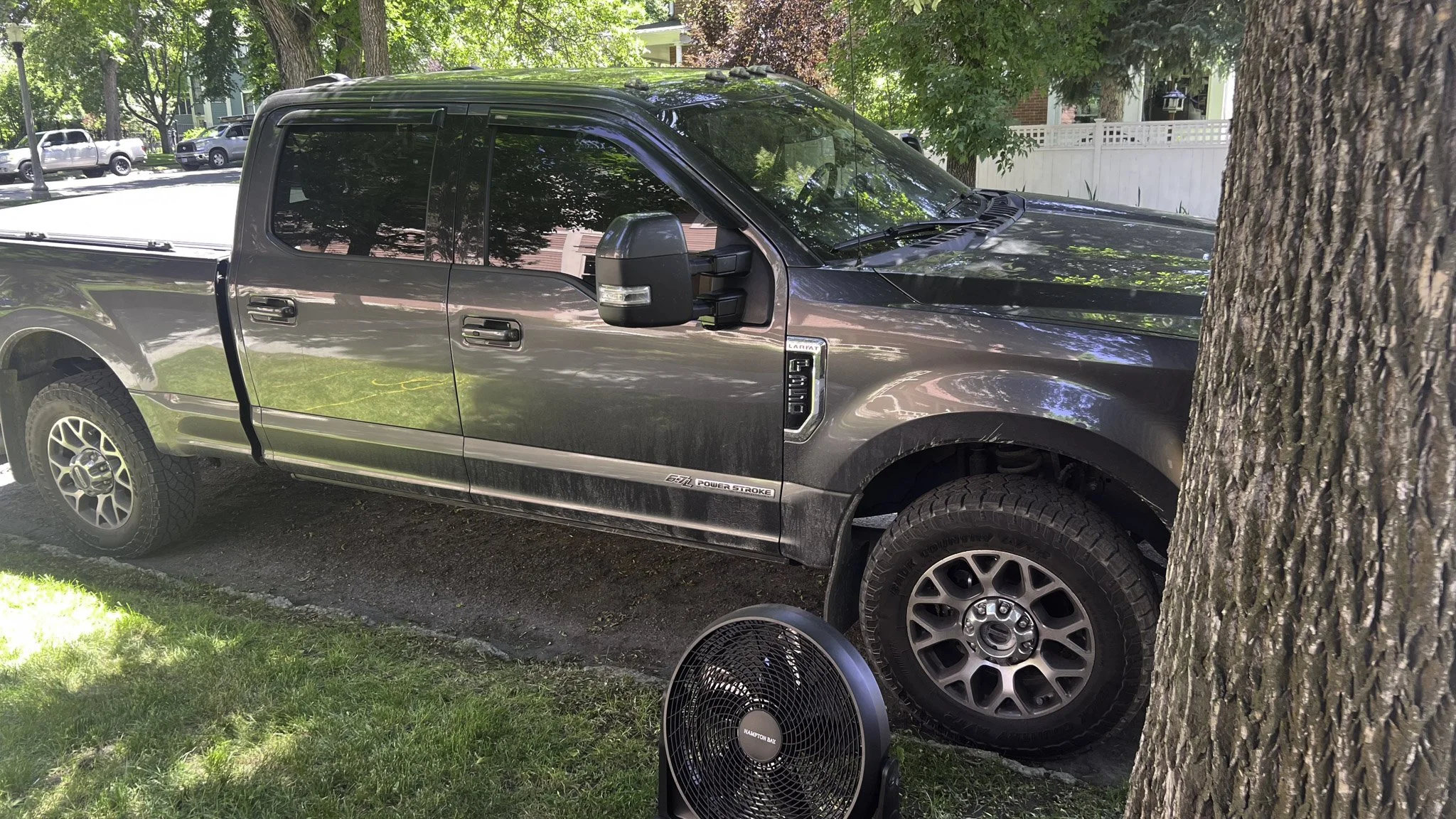 A gray pickup truck parked on a grassy area under trees with a fan sitting on the ground nearby. The truck is next to a tree, and there are other vehicles and houses visible in the background.