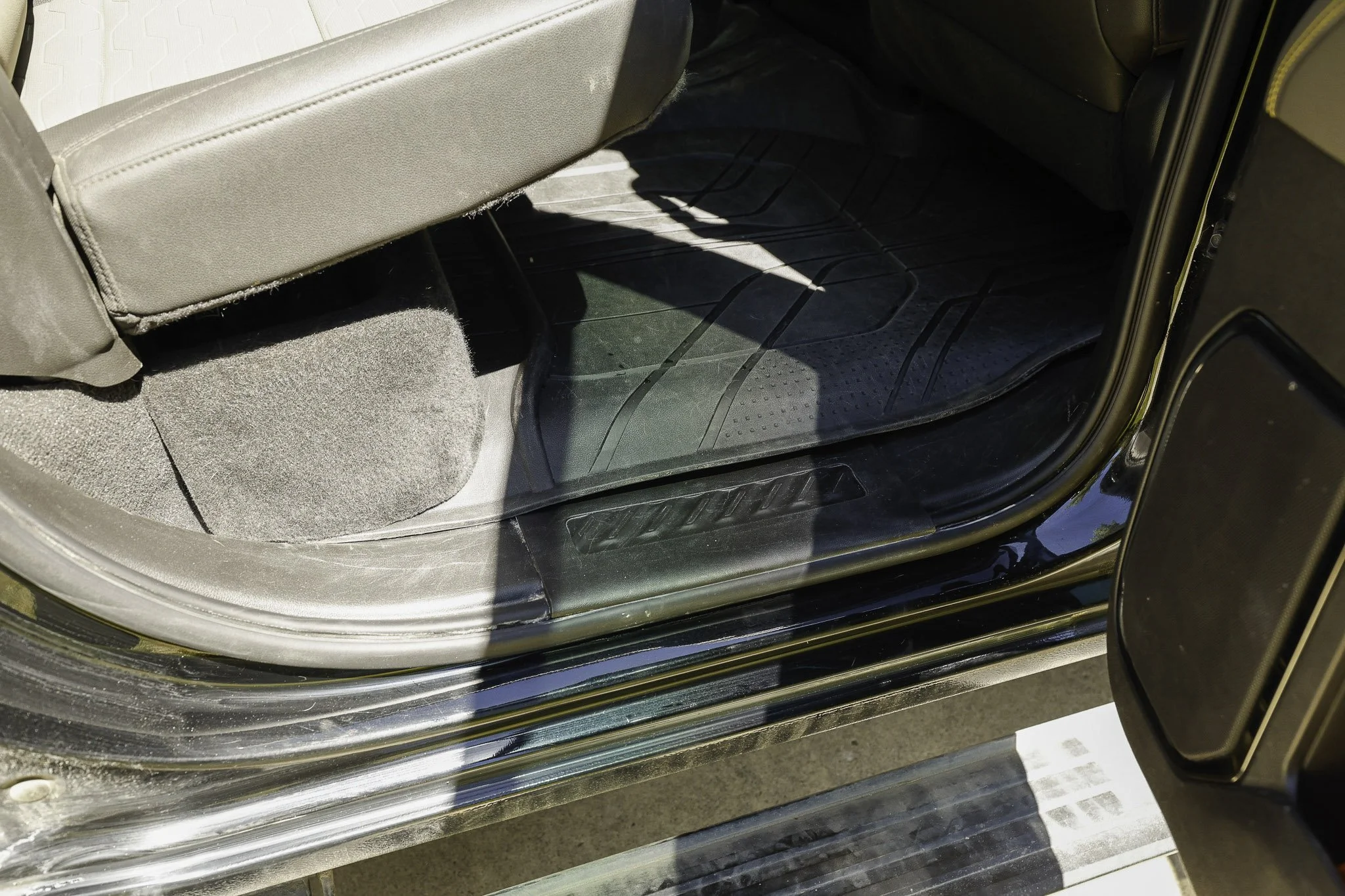 The image shows the interior of a vehicle, focusing on the front passenger side floor area. There is a black rubber floor mat, part of the vehicle's interior with some sunlight and shadow boundaries visible.