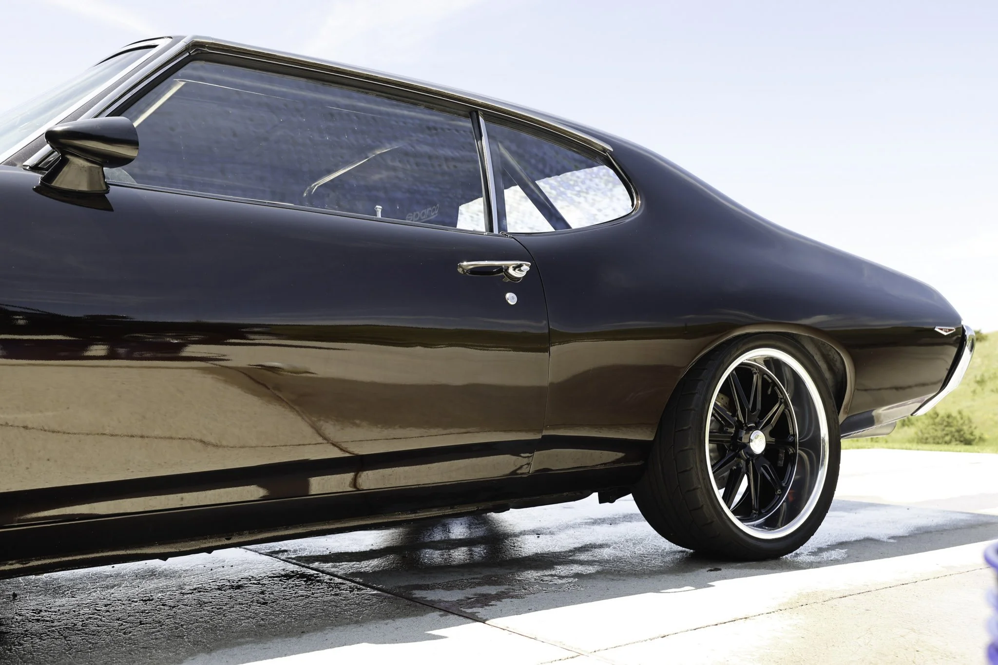Close-up of a vintage black sports car with black rims, parked on a concrete surface outdoors under clear blue sky.
