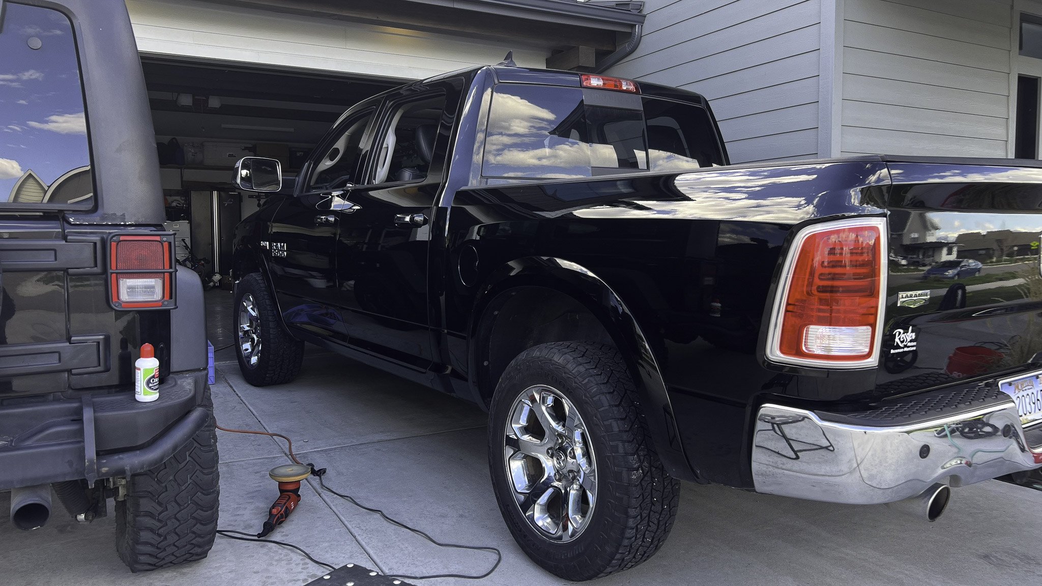 A black pickup truck parked in a garage, with a car detailing or washing process ongoing, indicated by a bottle of detailing product, a power buffer, and cables on the ground near the truck.