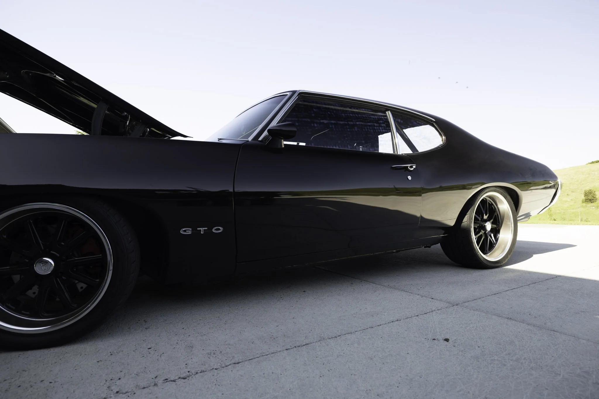 Side view of a black vintage muscle car with the hood open, parked on a concrete driveway with a grassy hill in the background.