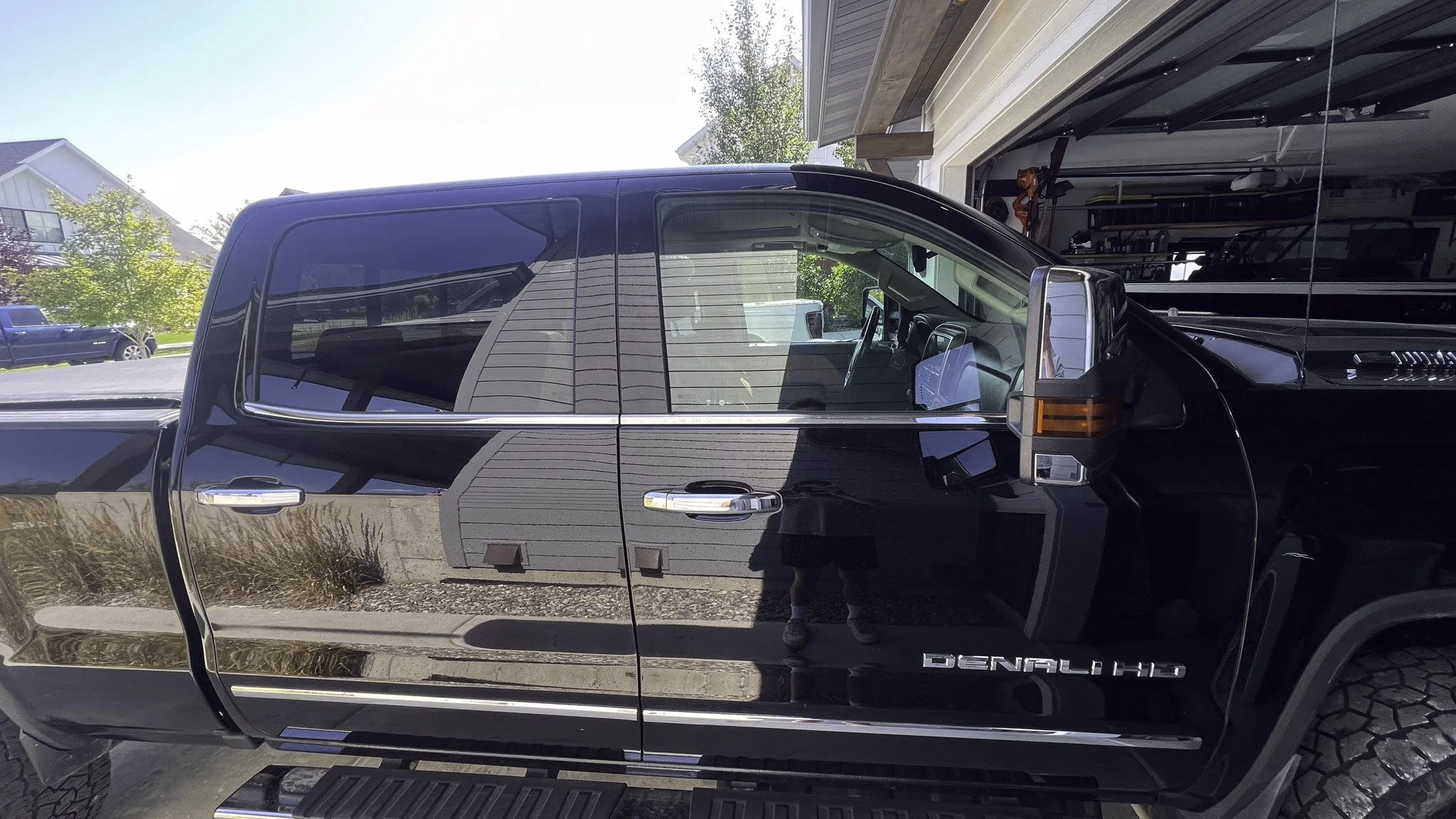 Black GMC Sierra Denali HD pickup truck parked in a driveway, with a garage open revealing a boat and tools inside. Reflection of the yard and person taking the photo visible on the truck's side.