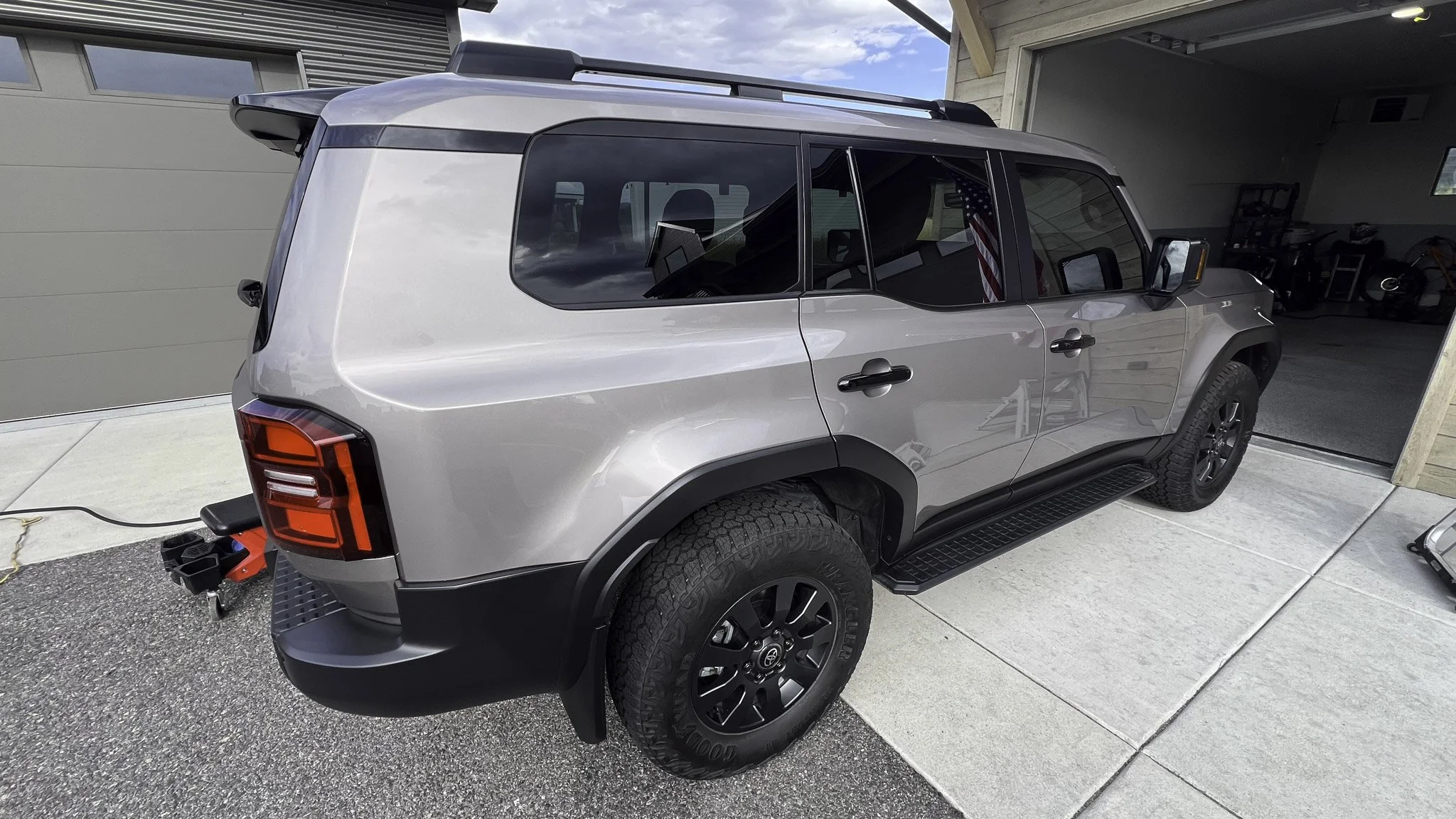 A silver and black Jeep parked in a driveway, with a garage in the background. The vehicle is attached to a red trailer hitch.