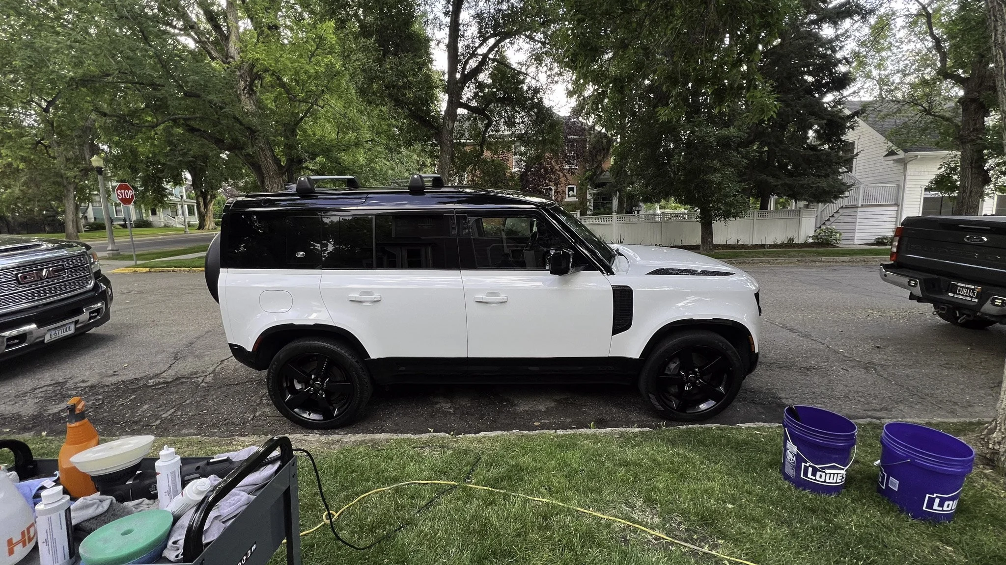 A white and black Land Rover SUV parked on a street with trees and houses in the background. There are two blue Lowe's buckets and maintenance supplies on the grass nearby.