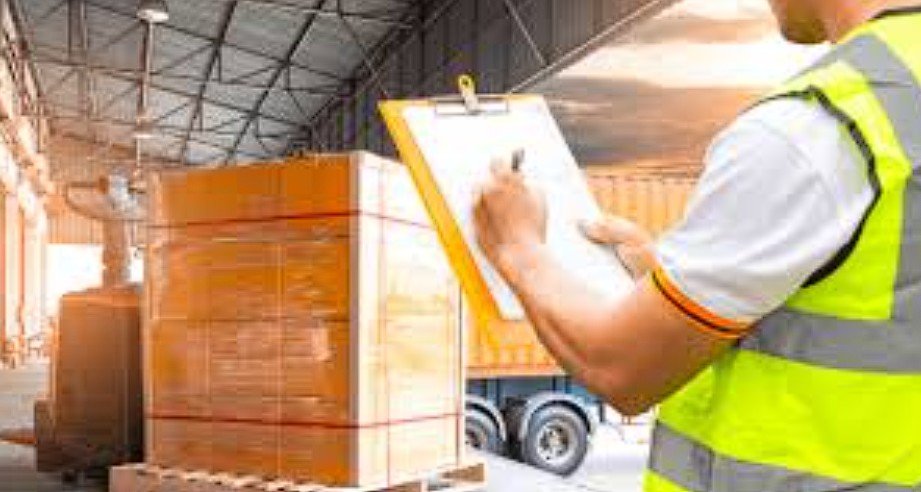 Worker in a safety vest writing on a clipboard in a warehouse with large wooden crates and a truck in the background.