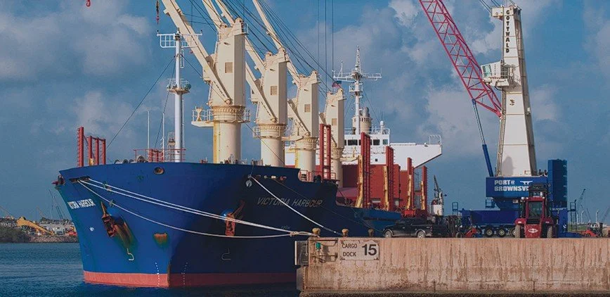 Cargo ship docked at port with cranes and trucks loading or unloading cargo, a sign indicating cargo dock 15, and a blue sky with clouds.