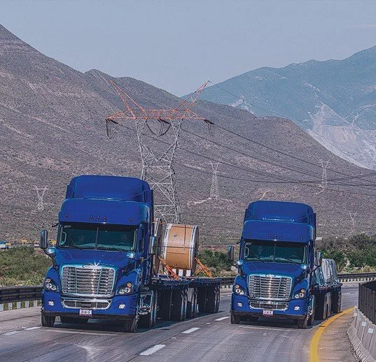 Two large blue semi-trucks towing a massive spool of cable on a highway with mountainous terrain and power lines in the background.