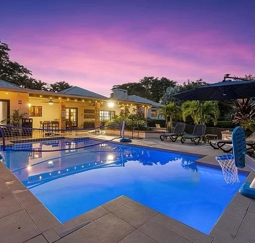Outdoor swimming pool area at sunset with lounge chairs, umbrellas, and a house in the background.