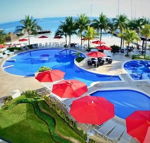 Swimming pool area with red umbrellas, lounge chairs, palm trees, overlooking the ocean.