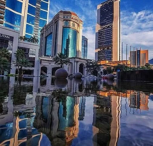 Cityscape with modern skyscrapers and a historic building reflected in a water feature in the foreground.