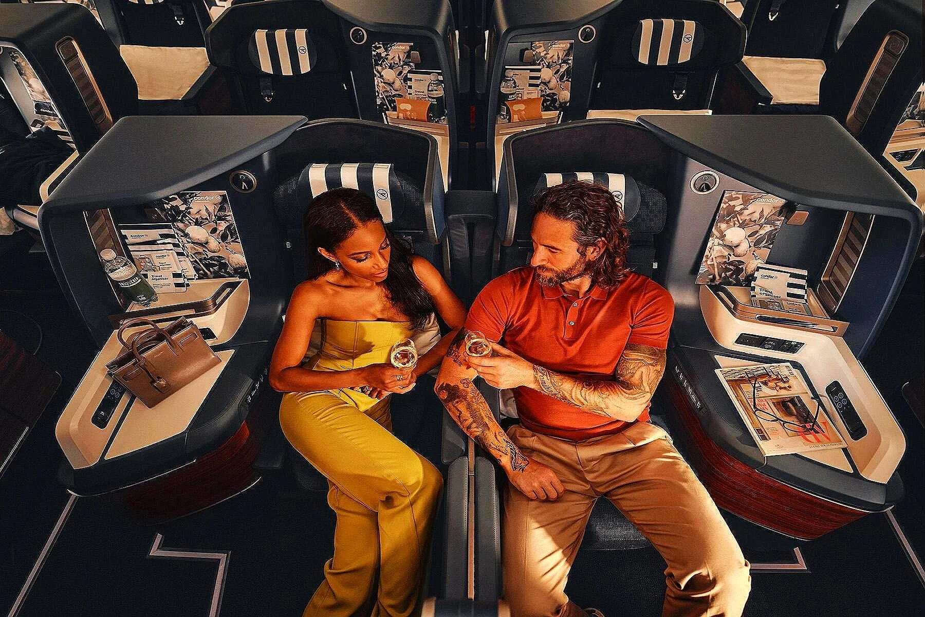 A man and woman in an airplane's private pod seats, facing each other and holding glasses of wine, engaging in conversation. The woman is dressed in a yellow outfit, and the man is wearing a red shirt with tattoos visible on his arms.
