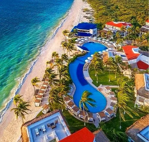 Aerial view of a beach resort with a curvy swimming pool, palm trees, sand beach, ocean, and resort buildings with colorful roofs.