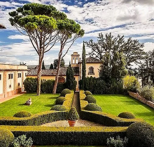 A manicured garden with trimmed hedges and topiary, tall trees, and historic buildings with Spanish-style terracotta roof tiles and a small bell tower in the background.