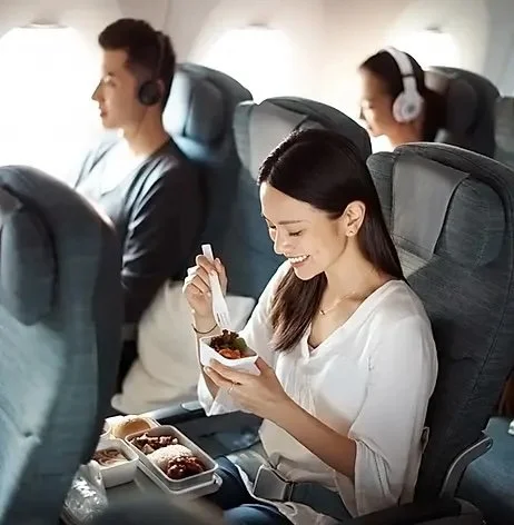 A woman smiling while eating a meal on an airplane with two other passengers in the background, all seated in airplane seats.