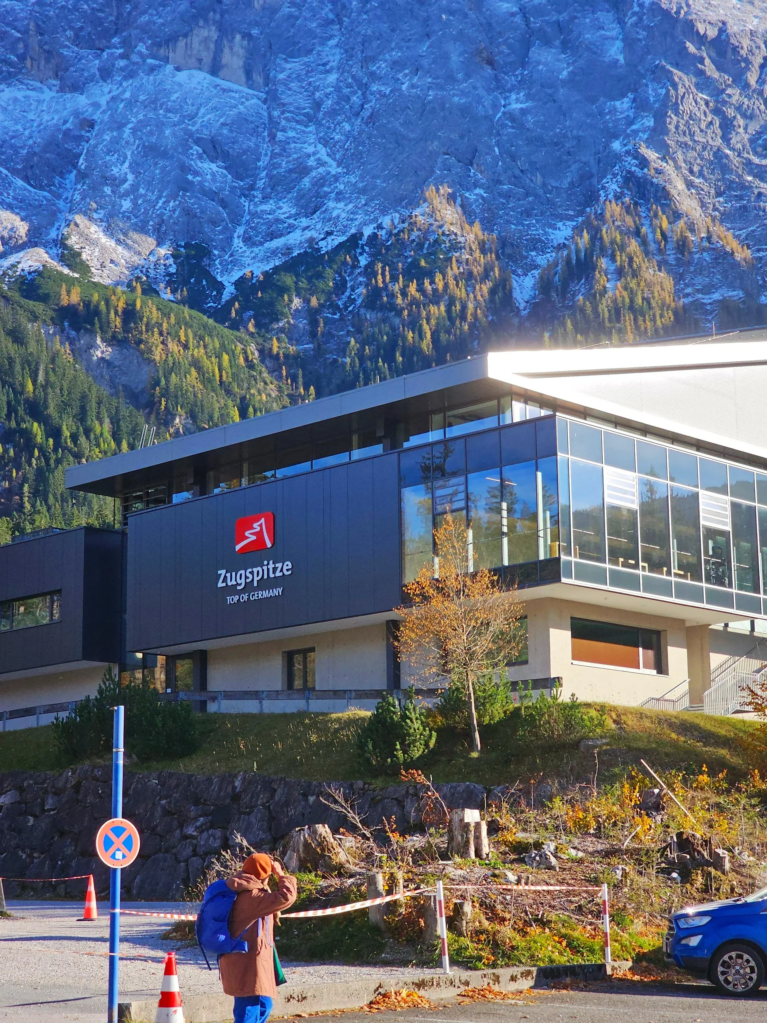 A modern building labeled 'Zugspitze TOP OF GERMANY' with trees and mountains in the background, and a person with a backpack in the foreground.