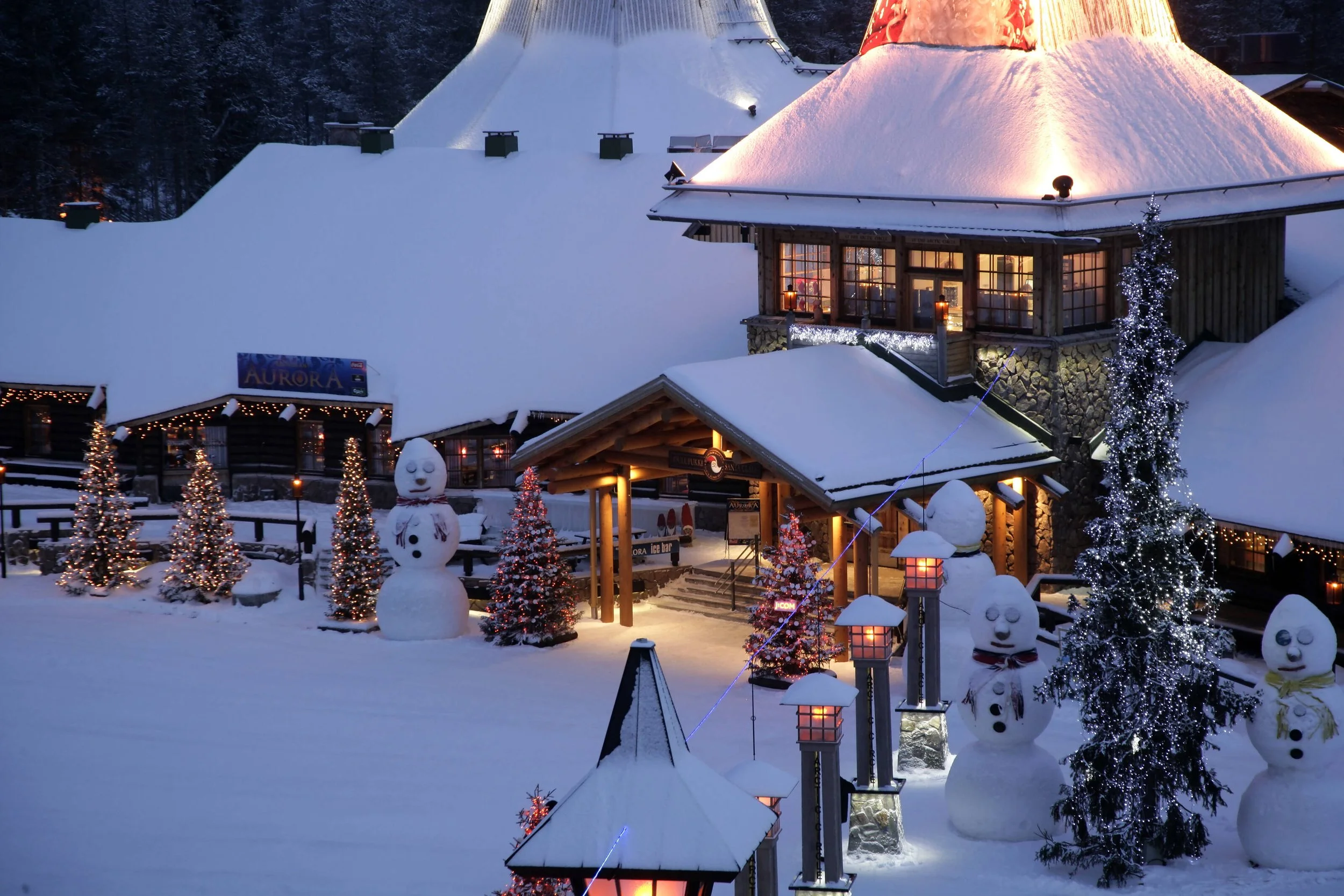 A snowy winter scene in front of a lodge decorated with Christmas lights. There are snowmen with scarves and buttons, small decorated Christmas trees, and tall trees wrapped in white lights. The lodge has large windows with warm light inside, and the area is illuminated by lamps with snow-covered roofs.