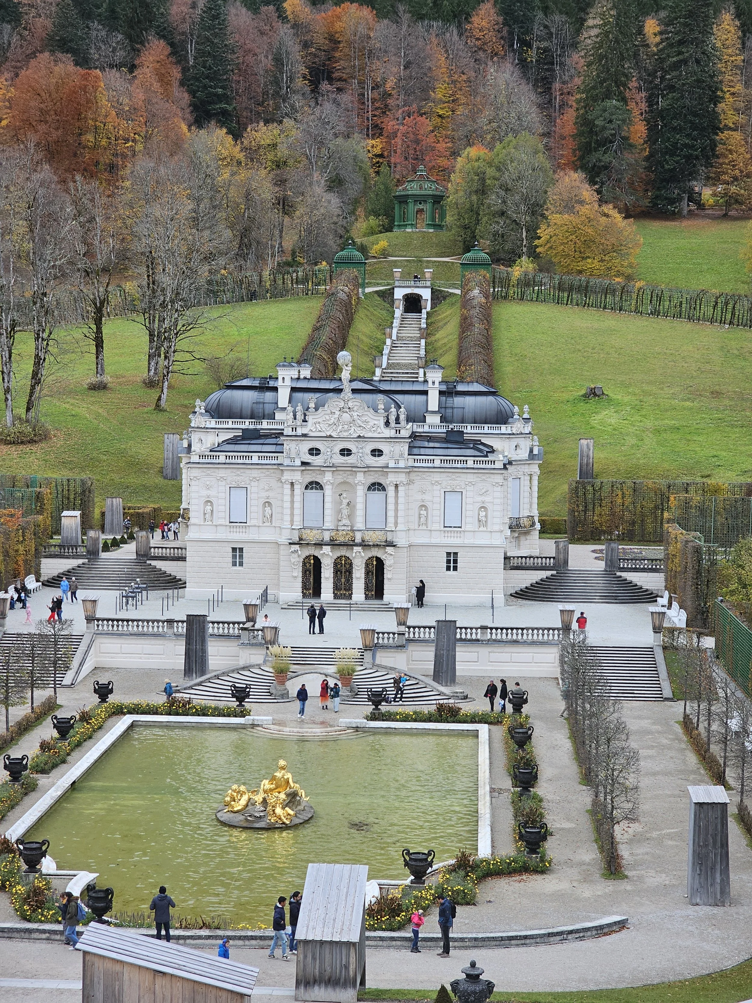 A grand white palace-style building with ornate details, a fountain with golden statues in front, and a large staircase leading up to a grassy hill with a green pavilion, set in a scenic park with trees in fall colors.