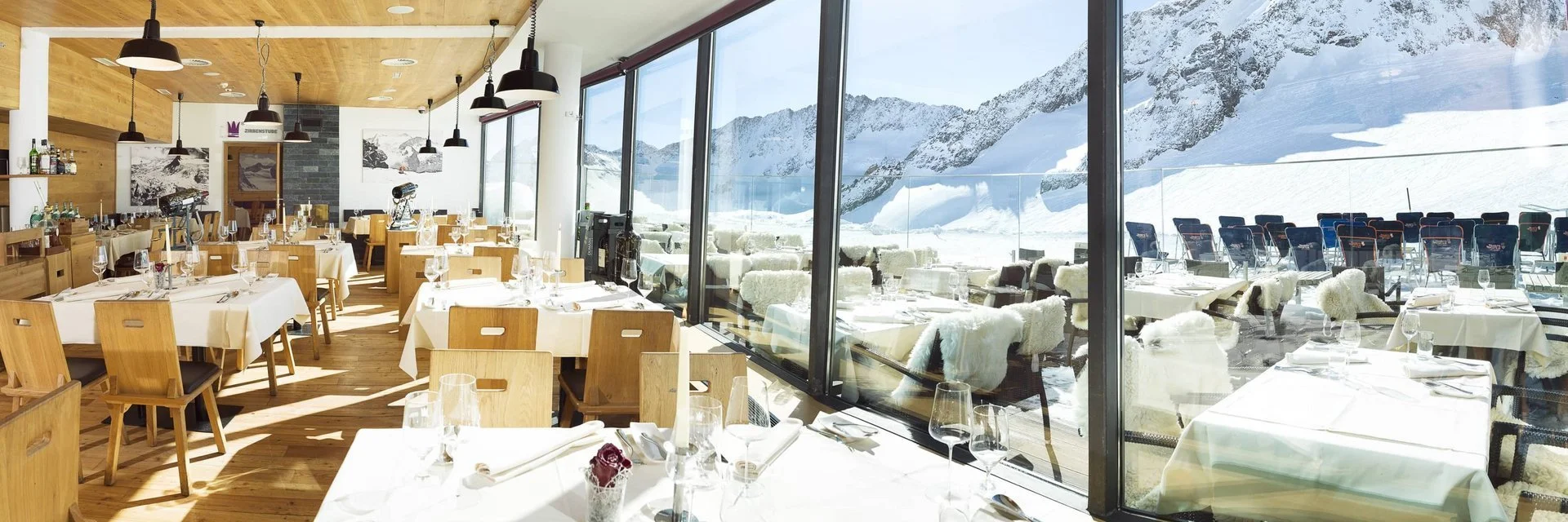Indoor and outdoor restaurant dining area with mountains and snow outside, tables set with glassware and white tablecloths.