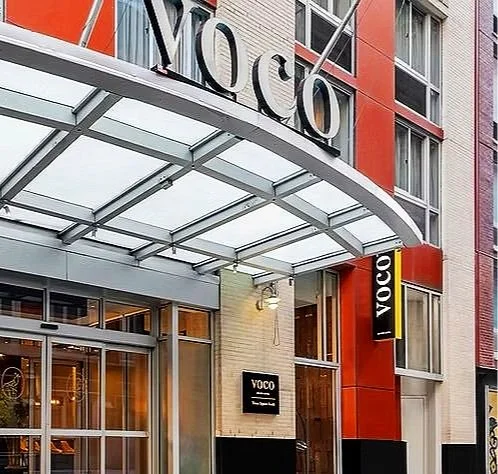 Exterior of a building with a large sign reading 'VOCO' above the entrance, featuring glass doors, a glass awning, and a modern design with red, white, and grey panels.