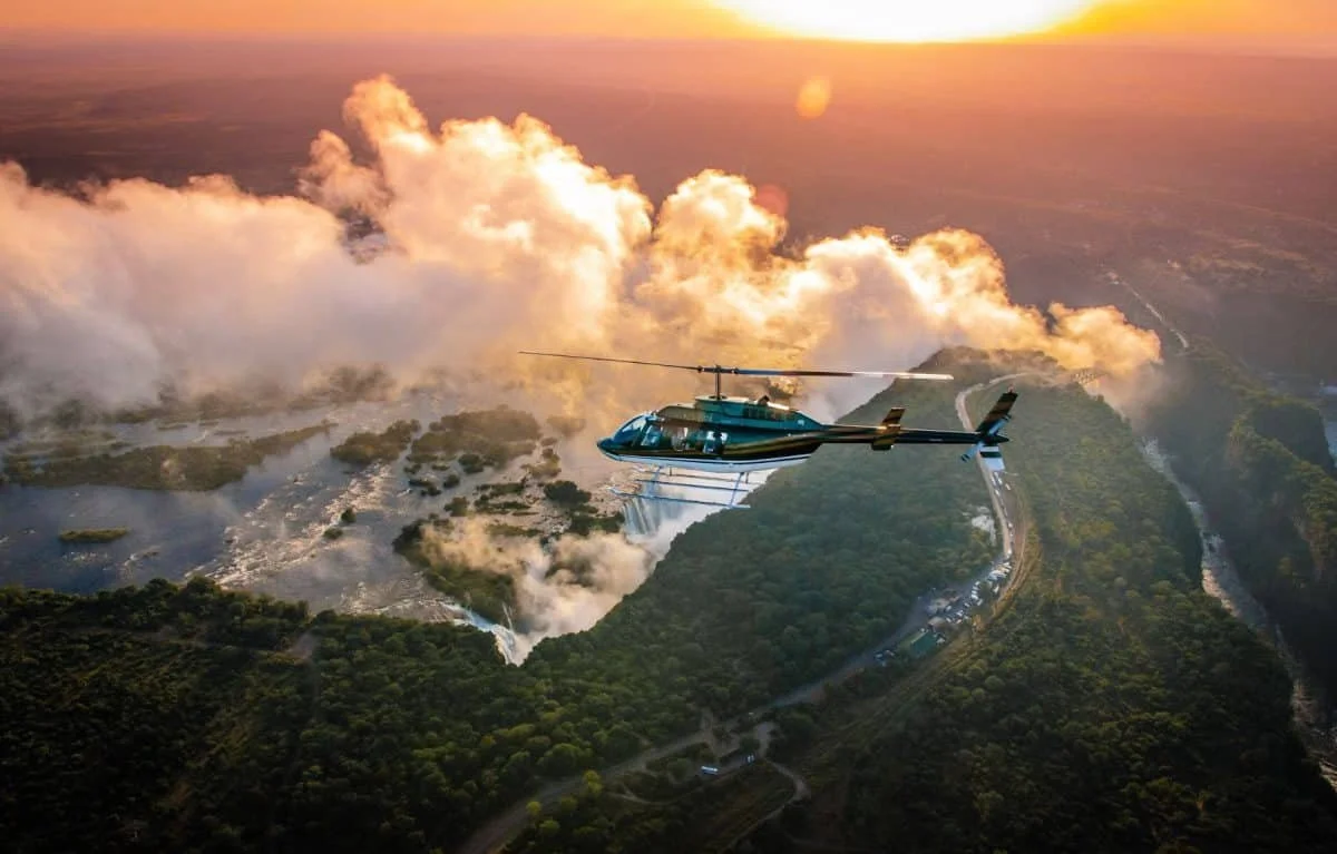A helicopter flying over a large river with waterfalls and lush green landscape during sunset.