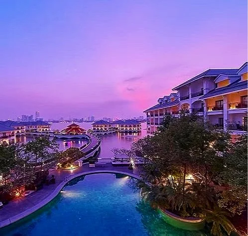 Sunset view of a luxurious waterfront resort with a swimming pool, modern buildings, and lush greenery, under a colorful sky.