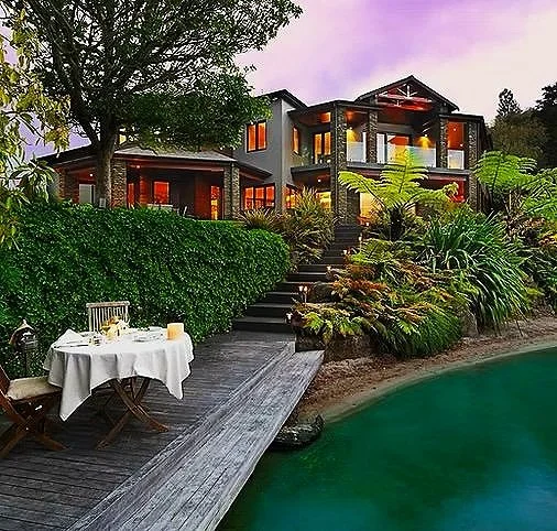 Luxury house with multiple levels and large windows, surrounded by lush greenery and plants, with an outdoor dining area on a deck next to a swimming pool at sunset.