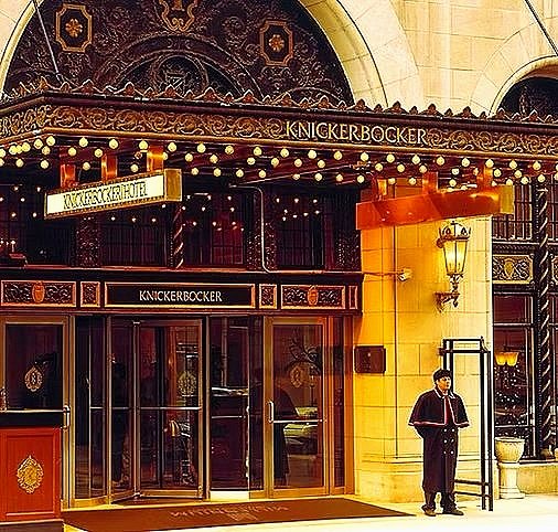 The entrance of Knickerbocker hotel with decorative marquee, warm lighting, and a doorman in uniform standing outside.