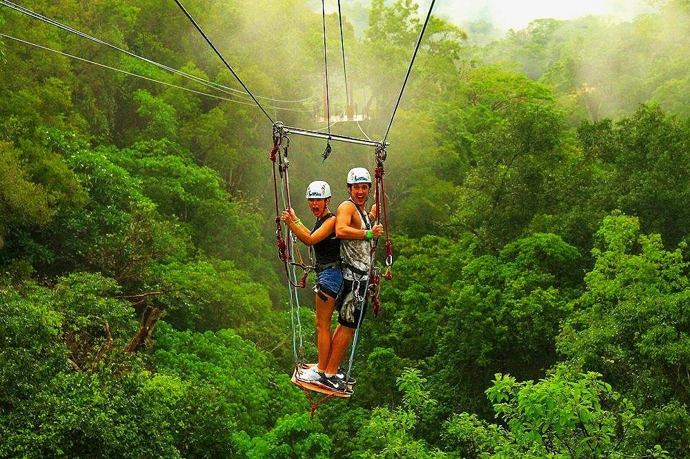 Two people riding a zipline over a dense green forest, both wearing safety helmets and harnesses.