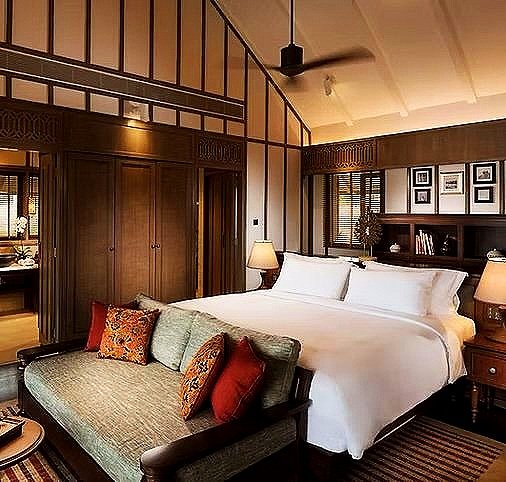 Hotel bedroom with a large bed, decorative pillows, bedside lamps, and a sitting area with a sofa, wood-paneled walls, and framed photos.
