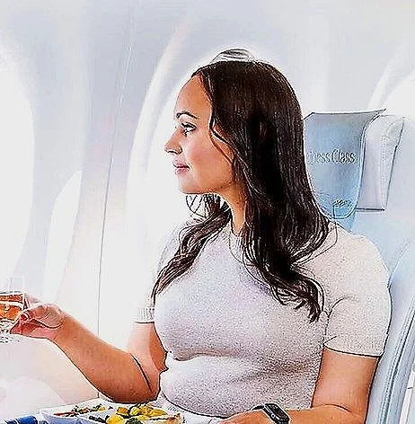 A woman sitting in an airplane seat, holding a glass of wine and looking out the window.