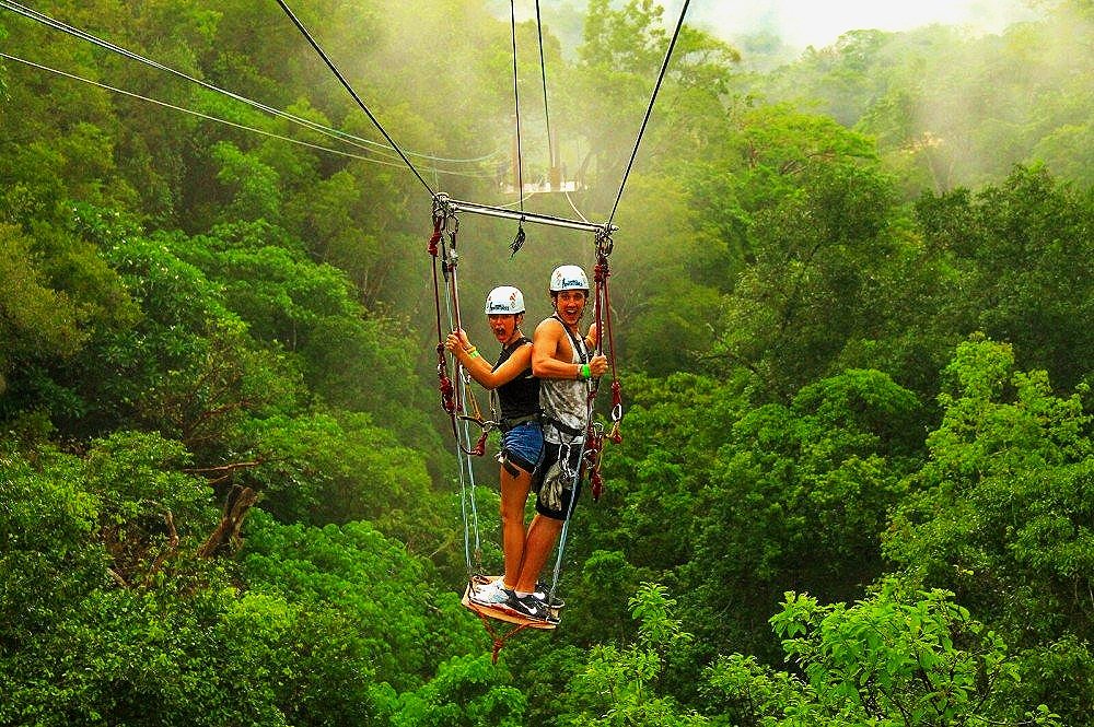Two people riding a zipline over a dense green forest, both wearing safety helmets and harnesses.