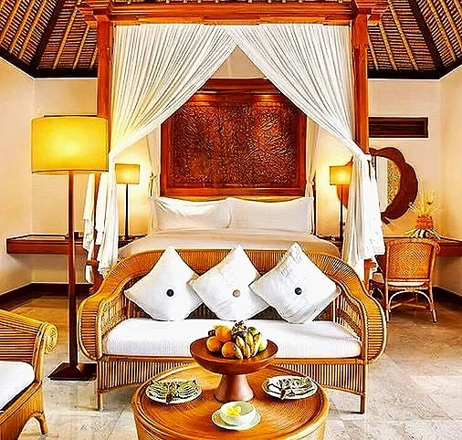 Hotel room with a canopy bed, wicker sofa with pillows, wooden side table with a fruit bowl, and a sitting area with a chair and mirror.