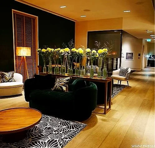 A modern hotel lobby with a black velvet sofa, white armchair, and a round wooden coffee table, decorated with a large arrangement of white flowers in glass vases, and patterned rugs on a wooden floor.