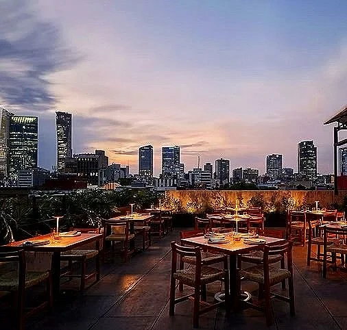 Empty rooftop restaurant with tables and chairs overlooking a city skyline at dusk.