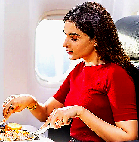 A woman with dark hair in a red shirt is eating a meal on an airplane