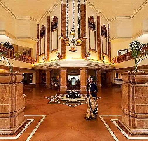 Elegant hotel lobby with a woman in traditional Indian attire walking across the polished wooden floor, grand architectural details, and tall windows with decorative drapes.