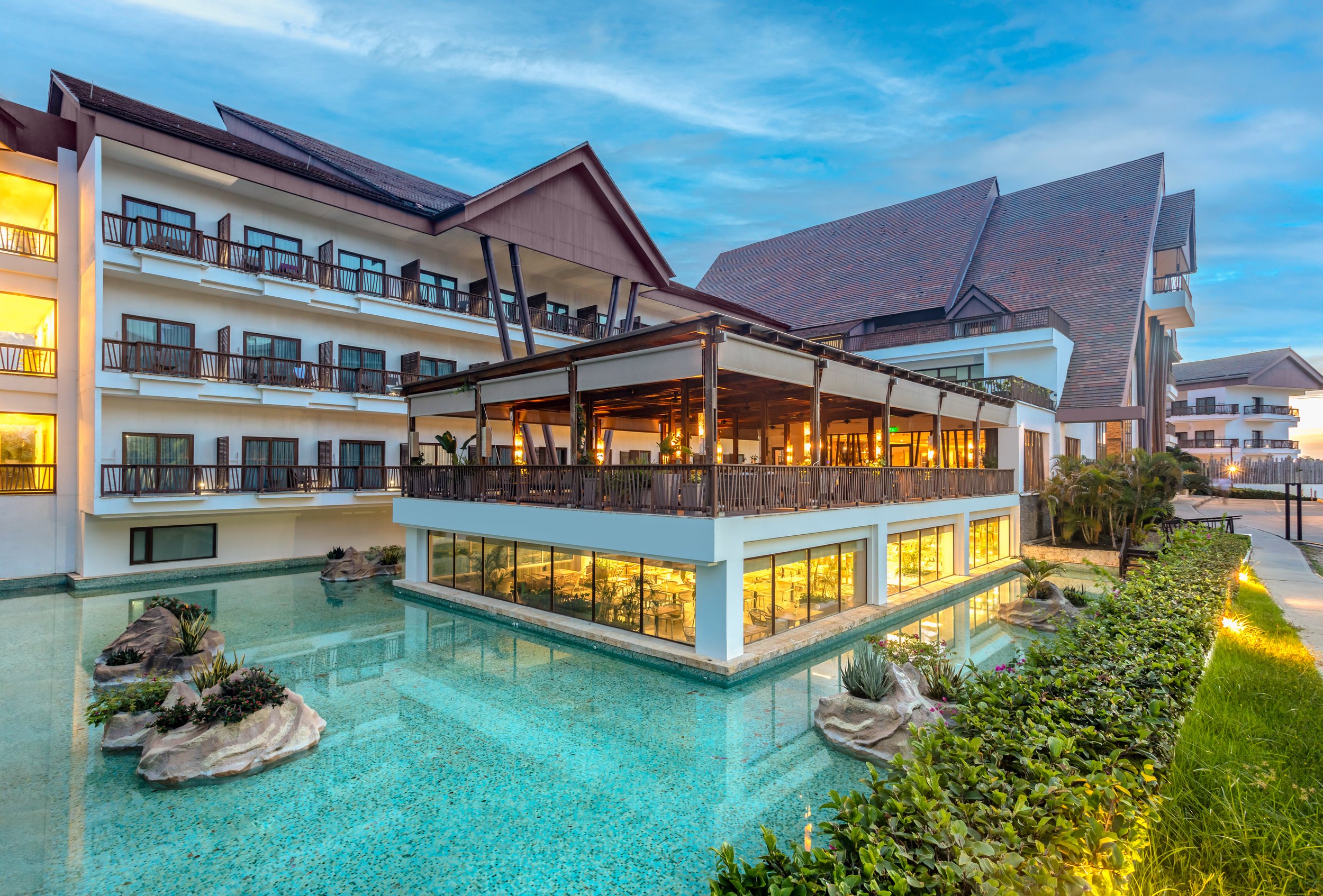 A modern hotel with multiple floors, balconies, and a large outdoor pool surrounded by rocks and greenery, illuminated by warm lighting at dusk.