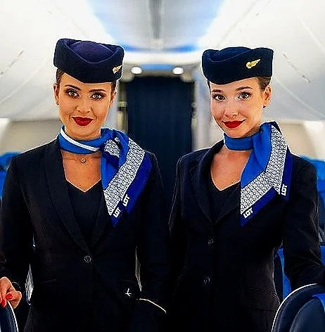 Two female flight attendants in uniform standing in an airplane cabin, wearing navy blue hats, matching scarves, and navy blazers.