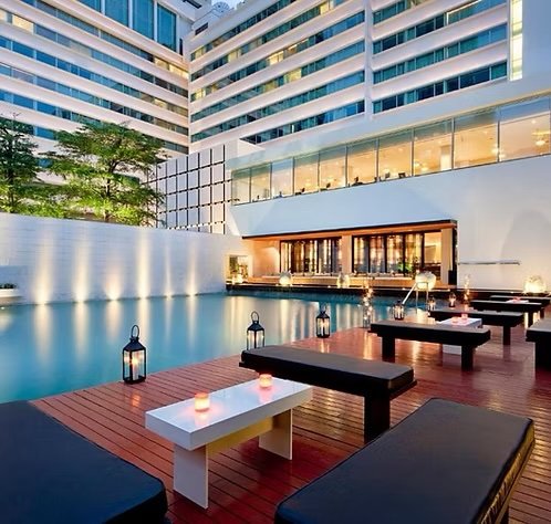 Modern hotel pool area with lounge chairs, lanterns, and candles at dusk, overlooking a cityscape with tall buildings.