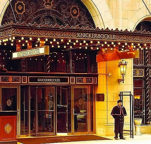 The entrance of Knickerbocker hotel with decorative marquee, warm lighting, and a doorman in uniform standing outside.