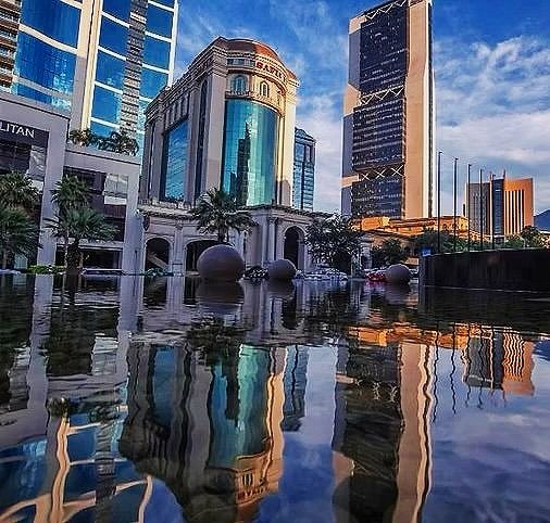 Cityscape with modern skyscrapers and a historic building reflected in a water feature in the foreground.