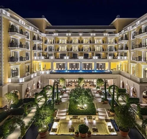 Night view of a lit-up multi-story hotel with balconies, a central garden with trees, and a swimming pool.