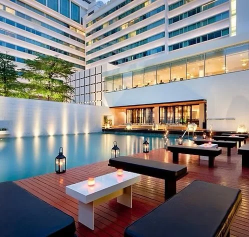 Modern hotel pool area with lounge chairs, lanterns, and candles at dusk, overlooking a cityscape with tall buildings.