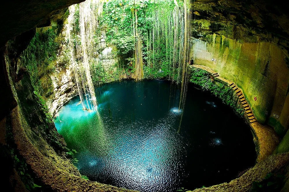 An underground sinkhole with a waterfall, surrounded by moss and greenery, leads to a deep, dark blue water pool with several small underwater lights illuminating the water.