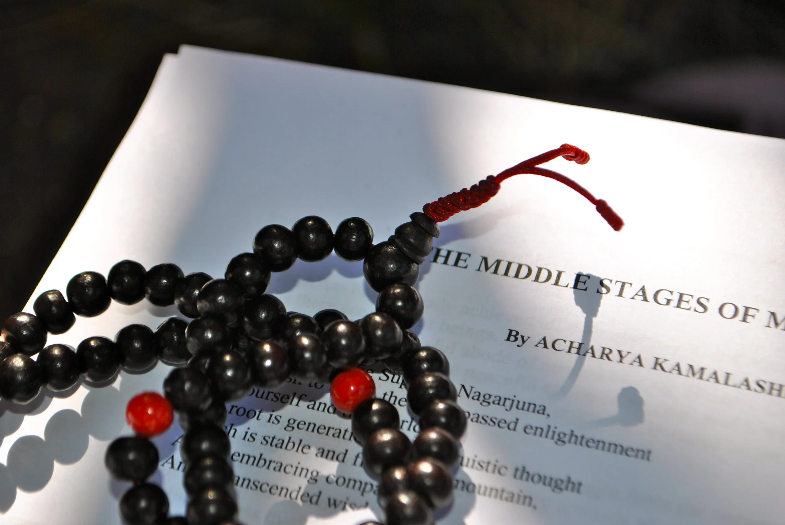 A black beaded mala necklace with red and orange beads resting on an open book titled 'The Middle Stages of M' by Acharya Kamalashe.
